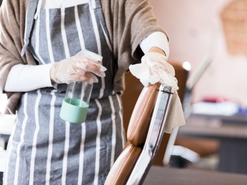 Osoba u kecelji i rukavicama briše naslon braon kožne stolice krpom i koristi sprej za čišćenje.