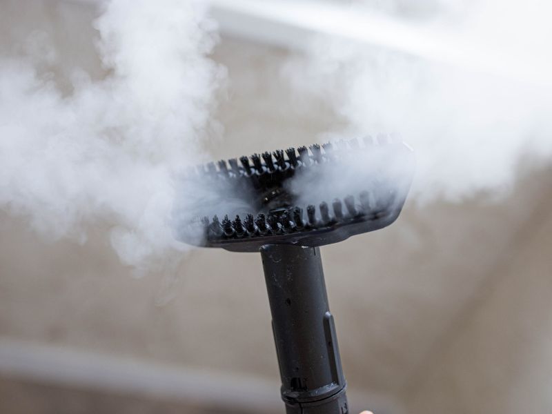 Paročistač sa crnom četkom ispušta paru tokom dezinfekcije tkanina i mrežastih površina.