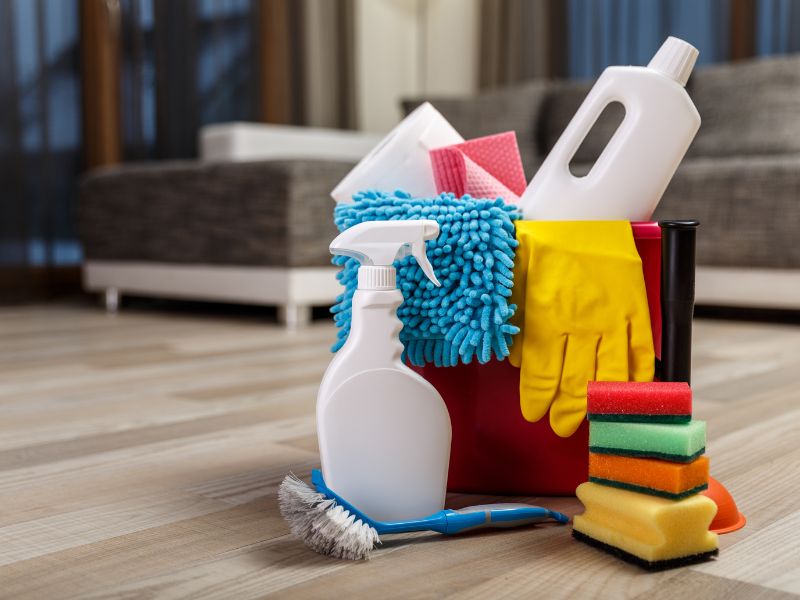 Kanta sa sredstvima za čišćenje, rukavicama, sunđerima, četkom i prskalicom postavljena na podu dnevne sobe.