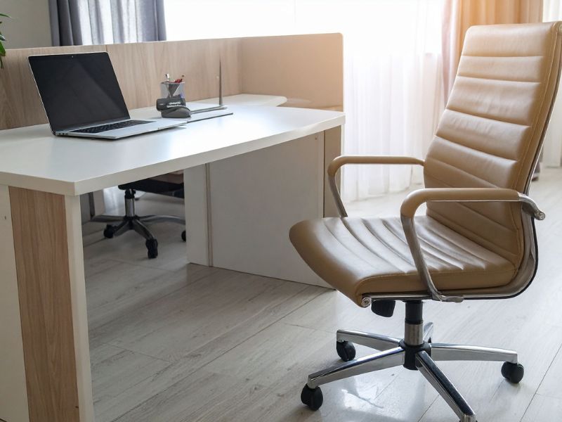 Bež kožna stolica sa metalnom bazom postavljena pored modernog radnog stola sa laptopom u osvetljenoj kancelariji.