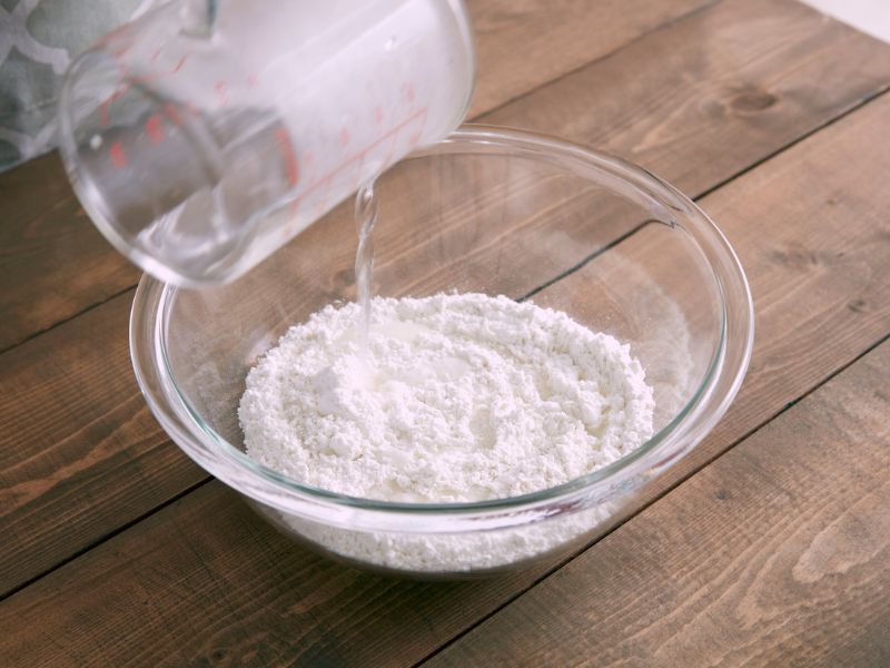 Staklena činija sa sodom bikarbonom u koju se sipa voda iz merne čaše, na drvenoj radnoj površini.