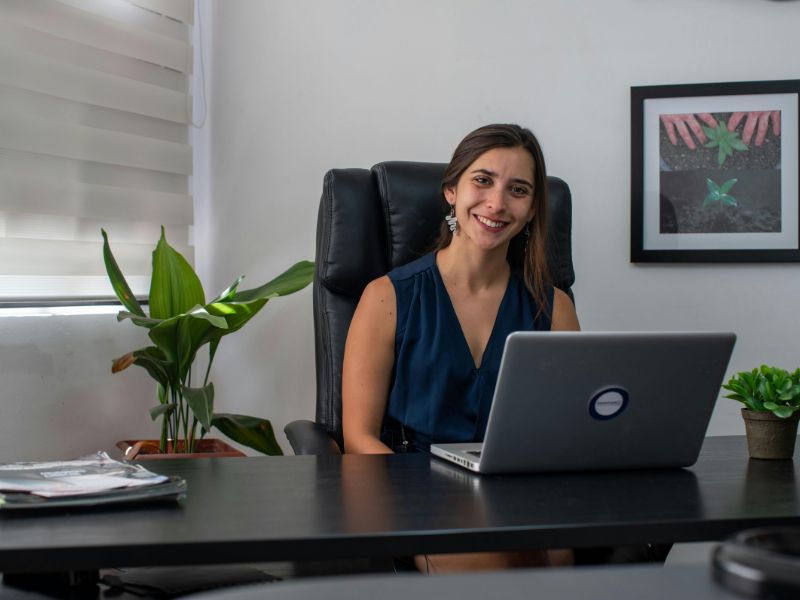 Žena sedi na crnoj kožnoj kancelarijskoj stolici za radnim stolom sa laptopom u modernoj kancelariji.