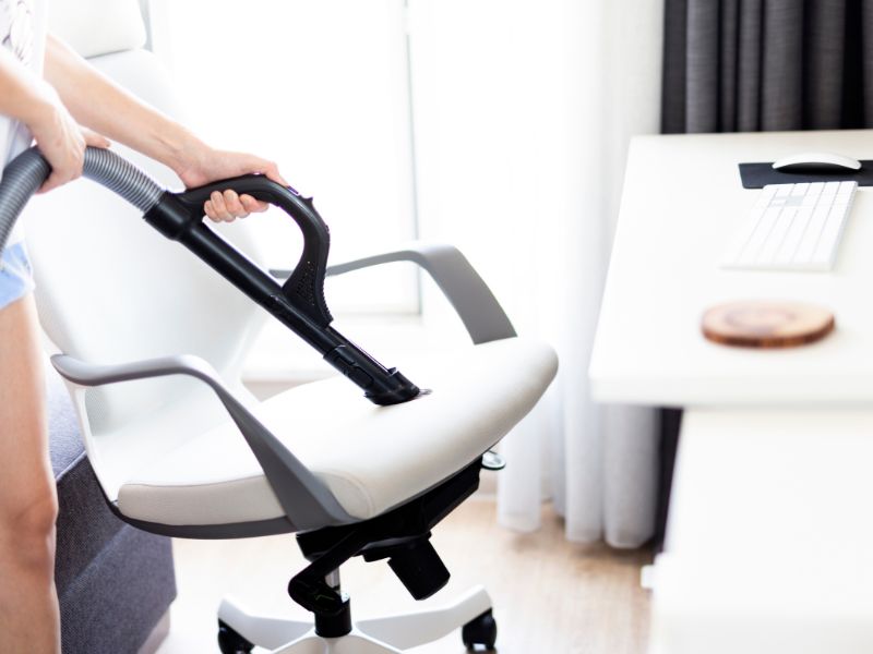 Osoba usisava belu kancelarijsku stolicu pomoću usisivača sa nastavkom za presvlake u svetloj radnoj sobi.