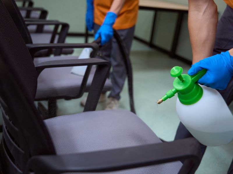 Radnici sa plavim rukavicama prskaju i usisavaju kancelarijske stolice sredstvom za čišćenje pomoću profesionalne opreme.