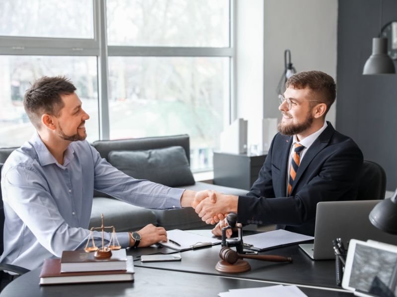 Pravnik i klijent se rukuju tokom sastanka u kancelariji, uz dokumenta, vagu pravde i druge pravne simbole na stolu.