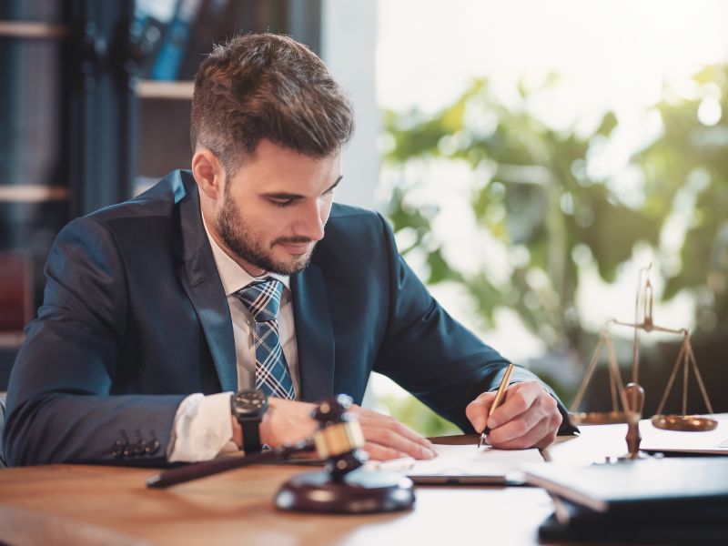 Advokat sedi za radnim stolom i potpisuje dokumente, okružen simbolima pravne profesije u mirnoj kancelariji.