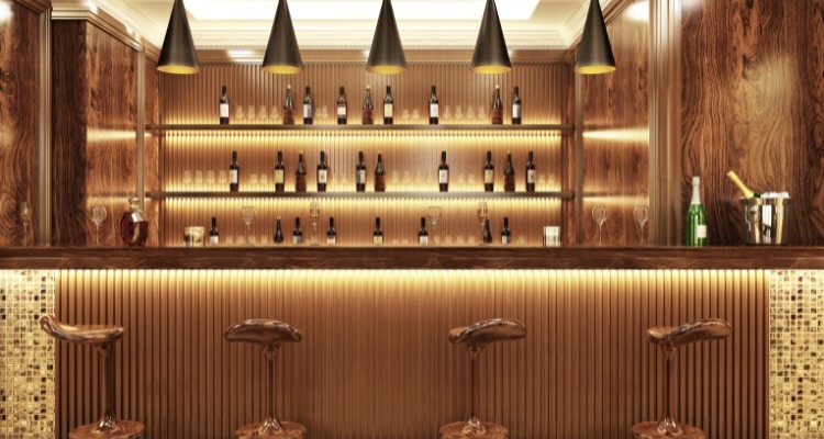 Bar sa drvenim oblogama, visećim lampama i policama uredno poređanim bocama, uz četiri elegantna barska stolice ispred šanka