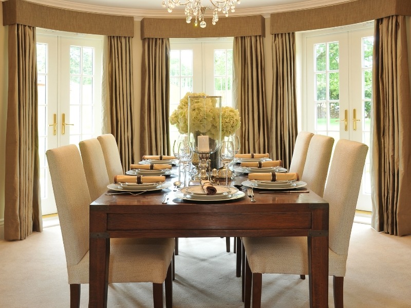 Elegantna trpezarija sa velikim drvenim stolom, osam tapaciranih stolica, cvetnim aranžmanom i velikim prozorima sa zavesama