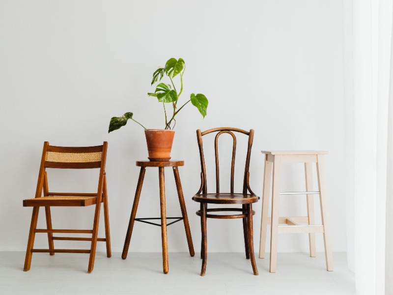 Tri različite drvene stolice i jedan visoki tabure pored kojih stoji saksija sa biljkom na malom stolu.