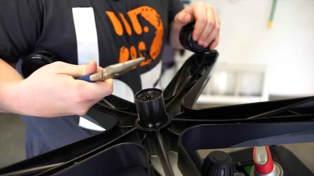 Osoba drži klešta dok radi na plastičnoj osnovi stolice sa uklonjenim točkićima