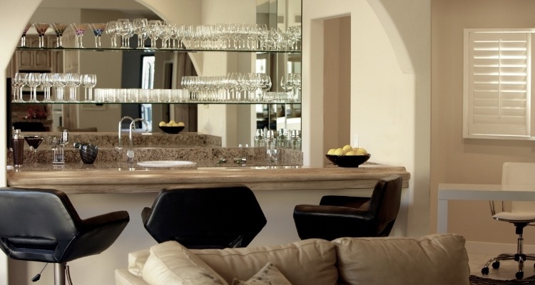 Svetao kućni bar sa mermernim pultom, ogledalima i policama punim čaša, uz tri moderne crne barske stolice.