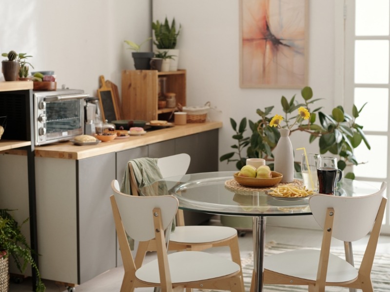 Moderna kuhinja sa drvenim elementima, okruglim staklenim stolom i belim stolicama, uz biljke i dekor na pultu.