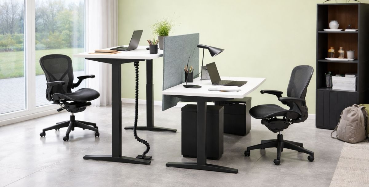 Moderan kancelarijski prostor sa dva podesiva radna stola, ergonomskim stolicama i svetlo zelenim zidom, osvetljen prirodnim svetlom.