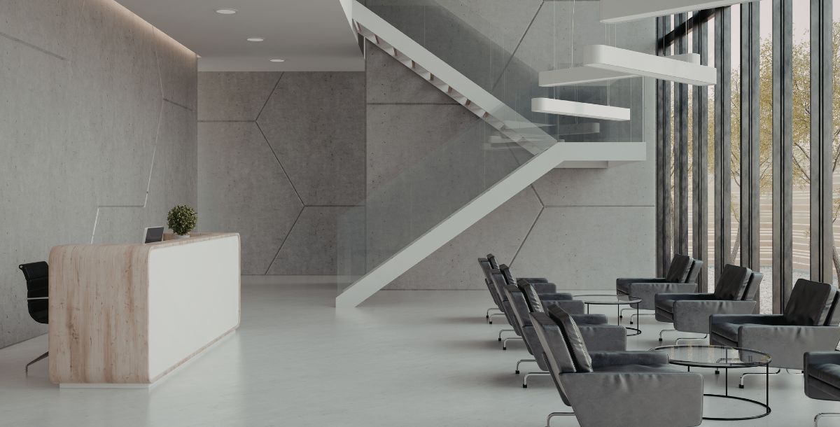 Moderna recepcija sa minimalističkim pultom, betonskim zidovima, staklenim stepeništem i prostorom za sedenje u savremenom poslovnom enterijeru.